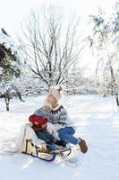 ung mor och henne söt liten son med retro kälke i en snöig parkera under solig dag foto