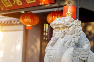 närbild och beskära kinesisk lejon statyett, sten skulptur i främre av kinesisk helgedom på suddigt kinesisk helgedom ingång med Sol blossa bakgrund. foto