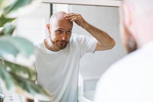 ung vuxen skäggig man ser i spegel i badrum rörande huvud orolig handla om handla om hår förlust foto