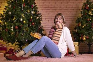 senior caucasian kvinna fira jul ensam i lycka och spänning medan innehav henne närvarande på Hem med dekorerad jul träd foto