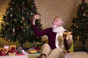 senior caucasian man fira jul ensam i lycka och spänning medan innehav hans närvarande på amning Hem med jul träd foto