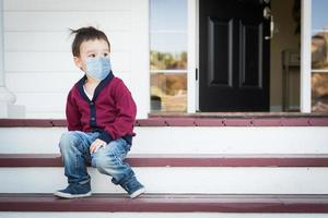 melankoli ensam blandad lopp pojke på främre veranda bär medicinsk ansikte mask under de coronavirus pandemi foto