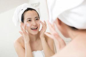 Lycklig ung asiatisk kvinna applicering ansikte lotioner medan bär en handduk och rörande henne ansikte i badrum foto