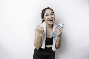 leende framgångsrik sportigt asiatisk kvinna Framställ med en handduk på henne axel och innehav en flaska av vatten foto