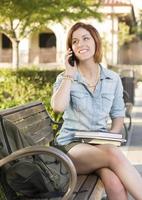 ung kvinna studerande utanför använder sig av cell telefon Sammanträde på bänk foto