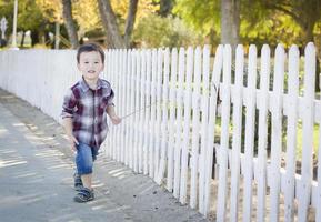ung blandad lopp pojke gående med pinne längs vit staket foto