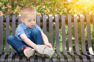ung pojke Sammanträde på en bänk sätta på hans skor på de parkera foto