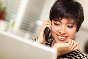 Söt leende multietnisk kvinna med cell telefon använder sig av bärbar dator foto