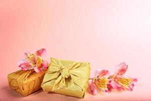 trendig insvept i gyllene textil- i furoshiki Metod gåva lådor och blommor alstroemeria på rosa. kopia Plats. foto