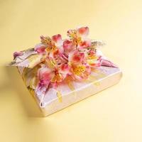 gåva låda trendig insvept i blommig tyg i japansk furoshiki Metod med blommor alstroemeria på gul bakgrund. foto