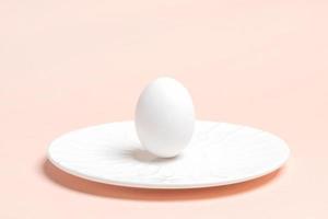 ett vit kyckling ägg är stående på vit tallrik på pastell rosa bakgrund. påsk minimal bild. foto