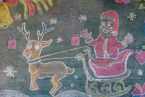 teckning av santa claus med ren på de svarta tavlan målad med annorlunda färger av krita förbi asiatisk studenter ritade dekorationer för de skolans jul firande. foto