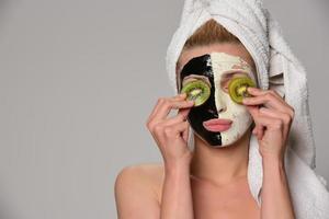 skön kvinna modell med svart och vit ansiktsbehandling kosmetisk mask innehav och bitande skivad orange. isolerat på grå bakgrund. foto