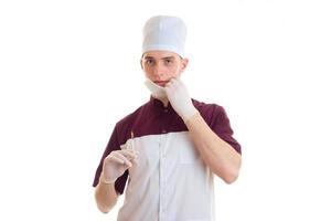 ung ärlig läkares hatt och med en mask på hans ansikte utseende hetero och innehav en spruta är isolerat på en vit bakgrund foto
