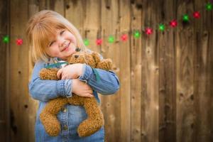 flicka innehav teddy Björn i främre av trädbevuxen bakgrund med jul lampor foto