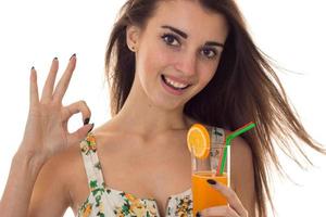ung Lycklig brunett flicka i sarafan med blommig mönster drycker orange cocktail leende på kamera och som visar ok isolerat på vit bakgrund foto