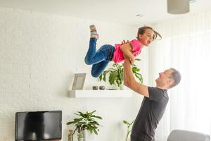 jag kan flyga. Lycklig pappa är lyft upp hans liten dotter medan stående i levande rum. foto