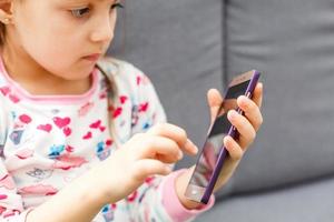 skön liten flicka spelar spel eller tittar på video på smartphone mobil. flicka tittar på tecknade serier eller bläddring internet, kopia Plats. sida se porträtt av liten flicka använder sig av smartphone medan Sammanträde. foto