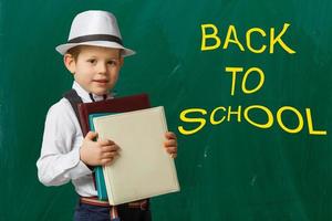 tillbaka till skola. rolig liten pojke i glasögon pekande upp på svarta tavlan. barn från elementärt skola med bok och väska. utbildning. foto