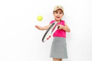 studio skott av en liten leende flicka klädd i rosa t-shirt och vit kjol - sportkläder för tennis på en vit bakgrund. hon står med en tennis racket och kastar en tennis boll foto
