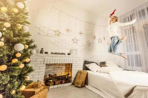 charmig flicka klädd i vit Tröja och byxor hoppar på de säng med grå filt och vit kuddar i en mysigt dekorerad rum med en ny år träd foto