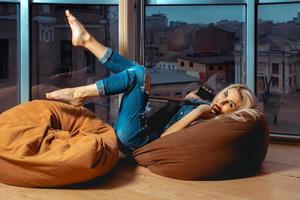 ung Söt blond kvinna Framställ på soffa i tillfällig jeans kläder nära stor fönster foto