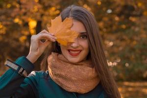 skön flicka med röd mun i de scarf innehar en lönn blad nära de ögon och ler foto