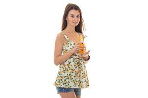 skönhet ung flicka i jeans shorts och sarafan med blommig mönster drycker orange cocktail och ser på de kamera isolerat på vit bakgrund foto