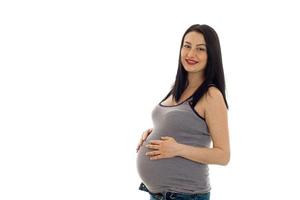 studio porträtt av ung gravid brunett kvinna i skjorta rörande henne mage och leende på kamera isolerat på vit bakgrund foto