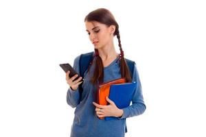 ledsen smart studerande flicka med ryggsäck på henne axlar och mapp för bärbara datorer i henne händer ser på mobil telefon isolerat på vit bakgrund foto