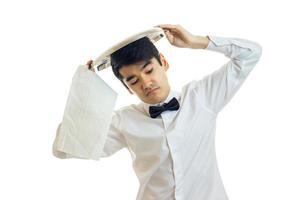 en ung servitör i en vit skjorta och svart rosett slips Uppfostrad gods bricka på hans huvud och stängd hans ögon foto