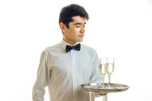 charmig ung servitör i en vit skjorta och med en fjäril innehav en bricka med två glasögon av champagne foto