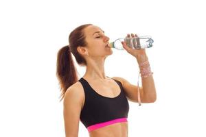 ung kondition flicka står sidled i de sporter topp och dricka vatten från en flaska foto