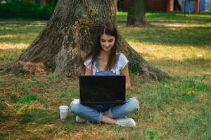 härlig ung flicka sitter på en gräs under en träd och använder sig av en lapto foto
