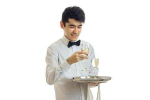 Lycklig ung man servitör i enhetlig med två glasögon av vin på silver- bricka leende foto