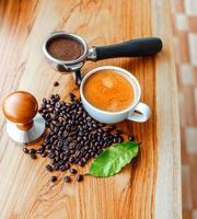 topp se av espresso kaffe kopp och Utrustning av Barista kaffe verktyg portafilter med manipulera och mörk rostad kaffe bönor med grön kaffe blad på trä- tabell foto