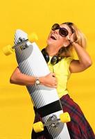 porträtt av Lycklig skön blond flicka håll henne trä- longboard skateboard på gul bakgrund vägg, se till de kamera och leende. urban scen, stad liv. hipster söt lady. foto