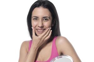 skön attraktiv brunett kvinna kontroll henne ansikte hud i främre av en spegel på vit bakgrund. foto