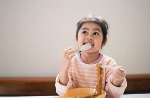 asiatisk bebis flicka njut av Lycklig använder sig av bestick sked och gaffel äter utsökt nudel i kök på dining tabell. Lycklig asiatisk bebis flicka öva äter förbi henne själv på dining tabell. bebis mat begrepp foto