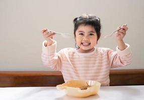 asiatisk bebis flicka njut av Lycklig använder sig av bestick sked och gaffel äter utsökt nudel i kök på dining tabell. Lycklig asiatisk bebis flicka öva äter förbi henne själv på dining tabell. bebis mat begrepp foto