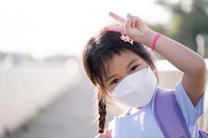 asiatisk barn flicka bär vit ansikte mask när gående till skola, skyddande mot damm, toxisk rök, luft förorening, ljus leende barn, v formad finger upp, unge bärande lila väska, tömma Plats. foto