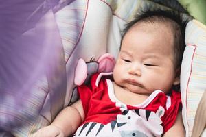 porträtt asiatisk bebis pojke är sömnig, litet barn är liggande i bil sittplats. suddigt lila fläkt kraft är ljus och Häftigt. barn bär röd onesie. barn är 1 månad gammal, nästan 2 månader gammal. foto
