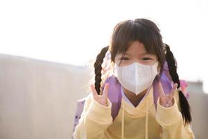 porträtt asiatisk söt flicka bär vit mask. barn log sött, innehav upp två fingrar i v form. förhindra luft förorening från toxisk damm. barn är bärande lila väska till skola.tom Plats. foto