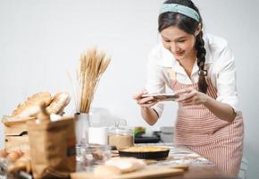 ung kvinna bär förkläde tar selfie Foto med hemlagad paj i kök. porträtt av skön asiatisk kvinna bakning efterrätt och har roligt tar Foto förbi smartphone för uppkopplad social. Hem kokkonst.