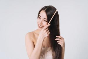 skönhet porträtt av Lycklig leende asiatisk kvinna med mörk lång hår kammande trä- hårkam på vit bakgrund isolerat foto