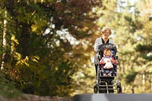 Lycklig senior lady tryckande hjul stol och barn. mormor och barn njuter en promenad i de parkera. barn stödjande Inaktiverad morförälder. familj besök. generationer kärlek och relation foto