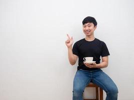 asiatisk man innehav kaffe kopp och punkt finger på Plats med Lycklig leende ansikte Sammanträde på stol på vit bakgrund foto