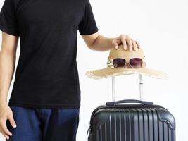 man svart skjorta stå med solglasögon hatt hängande ovan bagage på vit isolerad, resa semester begrepp foto