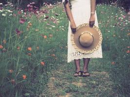 resande kvinna stående i blomma parkera och innehav hatt med skön utsikt, asiatisk kvinna resande vit klänning i blomma trädgård foto