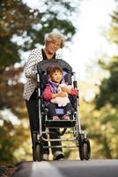 Lycklig senior lady tryckande hjul stol och barn. mormor och barn njuter en promenad i de parkera. barn stödjande Inaktiverad morförälder. familj besök. generationer kärlek och relation foto